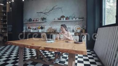 美丽的年轻女子坐在厨房的木桌旁，喝茶或咖啡，在她的<strong>银色</strong>笔记本电脑上打<strong>字</strong>